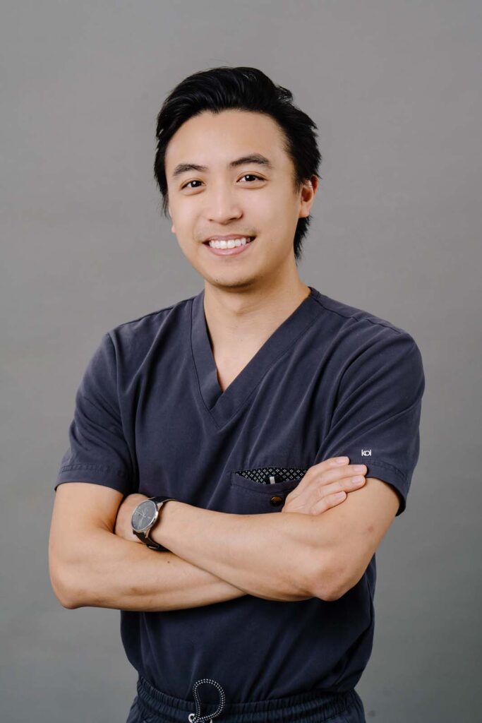 Hero shot of a dentist from Missouri City Dentistry crossing his arms and wearing a dark blue scrubs and a black wrist watch. The background is a plain gray color.