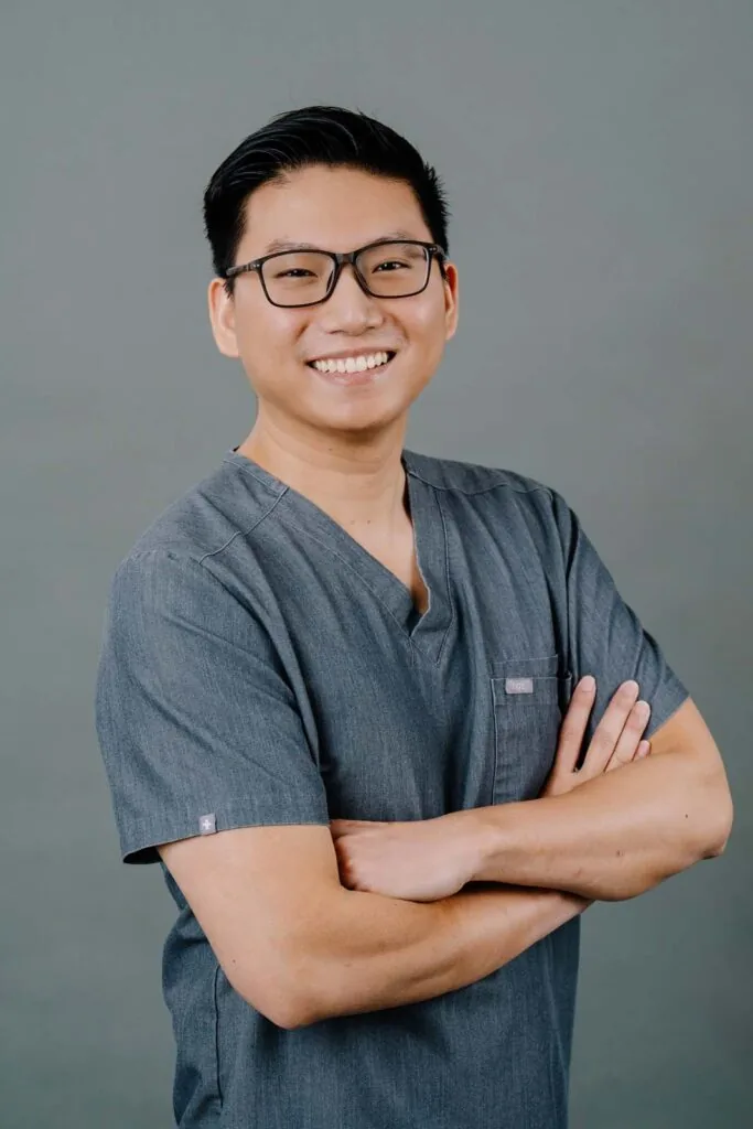 Hero shot of a dentist posing and wearing a dark gray scrubs while crossing his arms against a plain gray background.