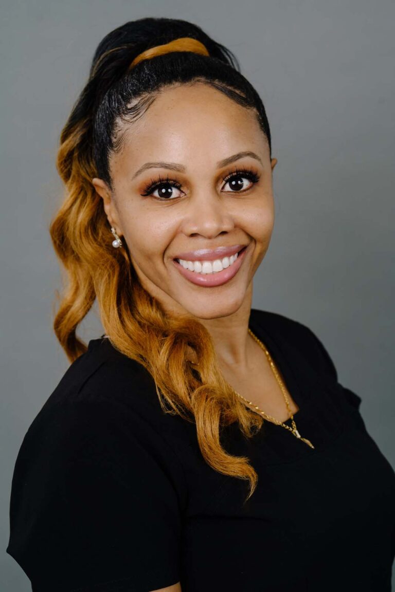 Head shot of a dentist with curly hair that transitions from black to blonde wears a black shirt and a gold necklace. The background is a plain gray backdrop.