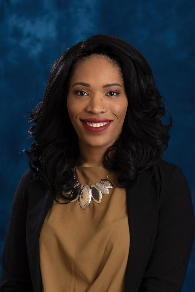 A person with long, curly black hair wears a black blazer over a mustard-colored top, with a necklace featuring three large, leaf-shaped pendants. The background is a textured blue backdrop.