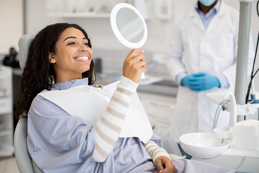 Smiling teen dental patient holding mirror after check‑up, dentist standing nearby in modern clinic.
